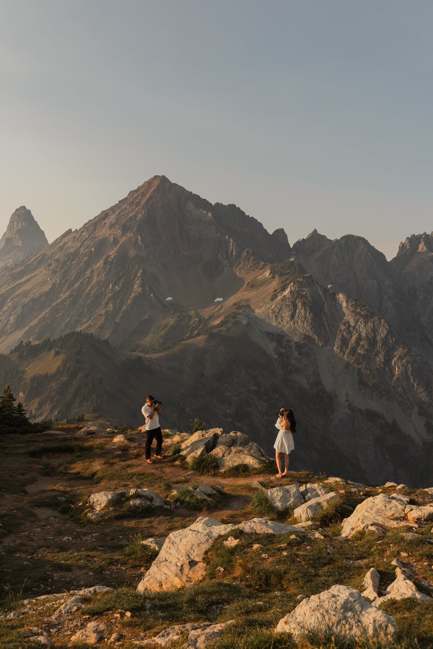 Two people photographing each other on a rocky ridge, with jagged mountain peaks behind them at golden hour