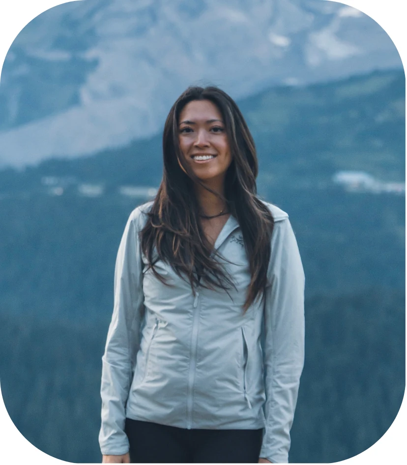 Angela smiling outdoors, with mountains and forest behind her