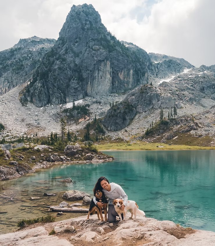 Angela with her two dogs at the edge of a turquoise alpine lake, with a rocky peak behind them